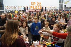 Tłum uczestników „Mikołajkowego Kiermaszu Rozmaitości” na forum Szkoły Podstawowej nr 22 w Płocku. W tle widnieje duży napis „SP 22 PŁOCK”, a na pierwszym planie dzieci i dorośli przeglądają stoiska z zabawkami, rękodziełem i biżuterią.