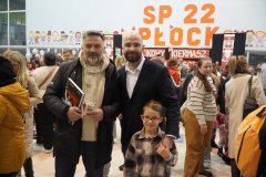 Wiceprezydent Płocka oraz trener piłki ręcznej Wisły Płock pozują do zdjęcia z chłopcem podczas mikołajkowego kiermaszu. Jeden z mężczyzn trzyma książkę o tematyce sportowej. W tle widoczny jest tłum uczestników oraz duży napis na ścianie „SP 22 PŁOCK” i dekoracje z dziadkami do orzechów.