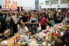 Tłum uczestników „Mikołajkowego Kiermaszu Rozmaitości” na forum Szkoły Podstawowej nr 22 w Płocku. W tle widnieje duży napis „SP 22 PŁOCK”, a na pierwszym planie dzieci i dorośli przeglądają stoiska z zabawkami, rękodziełem i biżuterią.