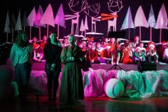 Na scenie znajduje się duża grupa dzieci i młodzieży, ubranych w świąteczne stroje. W tle dekoracje zimowo-bożonarodzeniowe: stylizowane choinki, białe elementy przypominające śnieg oraz duże sylwetki reniferów. Scena jest oświetlona kolorowymi światłami (różowe, zielone). Na pierwszym planie stoją trzy dorosłe osoby, jedna z nich mówi do mikrofonu i czyta z kartki. Za nimi dzieci siedzą i stoją w kilku rzędach, część ma czapki Mikołaja. Całość wygląda jak szkolne lub lokalne przedstawienie świąteczne.