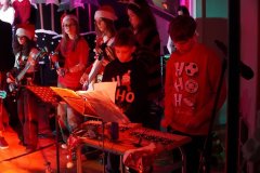 Na zdjęciu widać grupę młodych osób występujących na scenie podczas świątecznego koncertu. Część z nich gra na gitarach, a inni stoją przy stoliku z instrumentami perkusyjnymi. Wszyscy mają na sobie świąteczne swetry i czapki Mikołaja. Scena oświetlona jest ciepłym, czerwonym światłem, a w tle widać dekoracje w zimowym klimacie. Panuje radosna, świąteczna atmosfera.