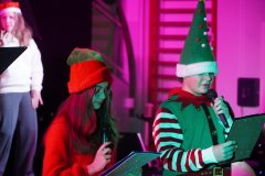 Na zdjęciu widać dwoje dzieci występujących na scenie podczas świątecznego przedstawienia. Oboje trzymają mikrofony i kartki z tekstem. Dziewczynka po lewej stronie ma długie włosy, czerwoną czapkę Mikołaja i czerwony sweter. Obok niej stoi chłopiec ubrany w zielony strój elfa z paskiem i w wysokiej zielonej czapce z białym futerkiem i czerwonymi pomponami. W tle widać osobę w białym swetrze i czerwonej czapce, a całą scenę oświetla różowo-fioletowe światło.