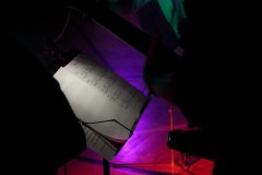 Na zdjęciu widać stojak z nutami oświetlony punktowym światłem. Obok znajduje się sylwetka muzyka trzymającego gitarę, widoczna w półcieniu. Tło jest rozmyte i oświetlone kolorowymi reflektorami w odcieniach fioletu, różu i czerwieni, co tworzy koncertowy nastrój.