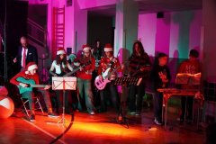 Na zdjęciu widać grupę młodych osób występujących na scenie podczas świątecznego koncertu. Kilkoro z nich gra na gitarach, a inni stoją przy instrumentach perkusyjnych. Wszyscy mają na sobie świąteczne ubrania – czerwone swetry, czapki Mikołaja i rogi renifera. Scena oświetlona jest kolorowymi światłami w odcieniach różu, czerwieni i zieleni. Po lewej stronie stoi dorosły mężczyzna w garniturze, obserwujący występ. Całość odbywa się w sali gimnastycznej, a atmosfera jest radosna i świąteczna.