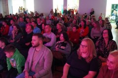 Na zdjęciu widać dużą grupę osób siedzących na krzesłach w sali podczas wydarzenia. Publiczność składa się z dorosłych i dzieci, wszyscy zwróceni są w stronę sceny, której nie widać w kadrze. Ludzie uśmiechają się i wyglądają na skupionych. Sala jest jasno oświetlona, z delikatnym różowo-fioletowym światłem padającym z przodu. W tle widać otwarte drzwi i korytarz, gdzie stoją kolejne osoby obserwujące wydarzenie. Atmosfera jest spokojna i przyjazna.