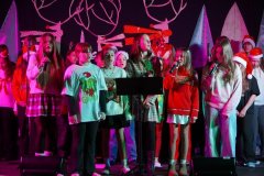 Na zdjęciu widać grupę dzieci i młodzieży występujących na scenie podczas świątecznego koncertu. Kilka osób stoi z przodu, trzymając mikrofony i śpiewając. Uczestnicy mają na sobie kolorowe, świąteczne ubrania – czerwone swetry, czapki Mikołaja, a niektórzy stroje z motywem reniferów i choinek. W tle widać dekoracje w kształcie białych choinek i dużych reniferowych poroży. Scena oświetlona jest kolorowymi światłami w odcieniach różu, czerwieni i zieleni, tworząc radosną, świąteczną atmosferę.