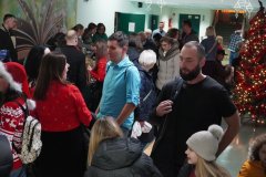 Na zdjęciu widać grupę ludzi zgromadzonych na korytarzu podczas świątecznego wydarzenia. W tle stoi ozdobiona choinka z kolorowymi światełkami. Wiele osób rozmawia ze sobą i uśmiecha się. Część uczestników ma na sobie świąteczne ubrania – czerwone swetry, czapki Mikołaja i zimowe kurtki. W głębi korytarza widać kolejne osoby. Atmosfera jest ciepła i radosna.