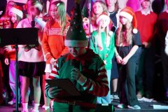 Na pierwszym planie dziecko przebrane za elfa (zielono-czerwony strój i wysoka czapka) trzyma mikrofon w jednej ręce i kartkę w drugiej, czytając tekst. W tle stoją inne dzieci i młodzież, również w świątecznych ubraniach: swetry, czapki Mikołaja, czerwone i zielone elementy. Po prawej stronie widoczny jest statyw z pulpitem na nuty. Oświetlenie sceny jest kolorowe, głównie różowo-zielone.