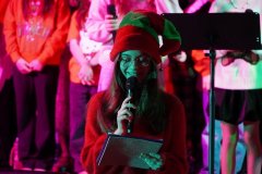 Na pierwszym planie dziewczynka w czerwonym stroju i czerwono-zielonej czapce elfa trzyma mikrofon oraz kartkę i czyta tekst. Ma okulary i lekko się uśmiecha. W tle stoją inne dzieci w świątecznych ubraniach. Obok niej widać statyw z pulpitem. Kolorowe światła sceniczne (różowe i zielone) oświetlają twarze i ubrania, podkreślając świąteczny klimat.