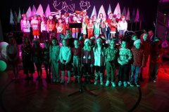 Na zdjęciu widać dużą grupę dzieci stojących na scenie. Dzieci ustawione są w kilku rzędach, najmłodsze z przodu, starsze z tyłu. Ubrane są w świąteczne stroje: swetry bożonarodzeniowe, sukienki, koszule oraz czapki Mikołaja. Na scenie znajduje się mikrofon na statywie. Tło sceny jest dekorowane w zimowym stylu – widać stylizowane choinki i renifery. Oświetlenie jest kolorowe, głównie różowe i zielone, co nadaje scenie uroczysty, świąteczny charakter.