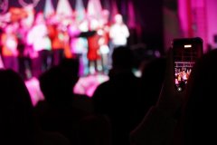 Zdjęcie wykonane jest z perspektywy widowni. Na pierwszym planie widać sylwetki osób siedzących lub stojących na sali. Jedna z osób trzyma w dłoni telefon komórkowy i nagrywa występ na scenie. Na ekranie telefonu widać dzieci występujące w świątecznych strojach. Sama scena w tle jest rozmyta i oświetlona intensywnymi, różowo-fioletowymi światłami.