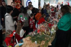 Zdjęcie przedstawia wnętrze szkoły, gdzie odbywa się świąteczny kiermasz. Przy długim stole stoją dzieci i dorośli. Na stole znajdują się świąteczne ozdoby: stroiki, koszyki, bombki, ręcznie robione dekoracje oraz drobne upominki. Dzieci ubrane są w czerwone i świąteczne stroje, niektóre mają opaski lub czapki Mikołaja. Atmosfera jest ruchliwa i radosna, w tle widać choinkę oraz wielu ludzi.