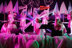 Na scenie występuje grupa dzieci przebranych w jasne, połyskujące kostiumy, przypominające anioły lub postacie baśniowe. Dzieci trzymają długie, półprzezroczyste wstęgi lub materiały, które rozciągają w różnych kierunkach, tworząc efekt promieni. Scena jest udekorowana zimowymi elementami, a oświetlenie jest różowe i fioletowe. Przed sceną widać statyw z nutami.