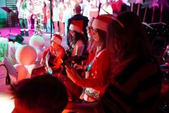 Na pierwszym planie widać dzieci siedzące lub stojące przy brzegu sceny. Dzieci ubrane są w czerwone stroje i czapki Mikołaja. Jedno z dzieci trzyma instrument muzyczny, prawdopodobnie gitarę lub ukulele. W tle widać inne dzieci stojące na scenie oraz mikrofony. Oświetlenie jest ciepłe, czerwone i różowe, co podkreśla świąteczny nastrój występu.