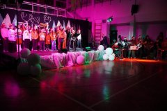 Początek formularza Dół formularza Na zdjęciu widać scenę udekorowaną w świątecznym stylu, ustawioną w sali gimnastycznej. Na scenie stoi grupa dzieci i młodzieży ubranych w czerwone i białe stroje, wiele osób ma na głowach czapki Mikołaja. W tle widać dekoracje przypominające zimowy las – białe choinki i reniferowe poroża. Przed sceną leżą duże białe kule przypominające śnieżne ozdoby. Po prawej stronie sceny siedzi kilku muzyków grających na instrumentach, oświetlonych kolorowymi światłami. Całość tworzy radosną, świąteczną atmosferę.