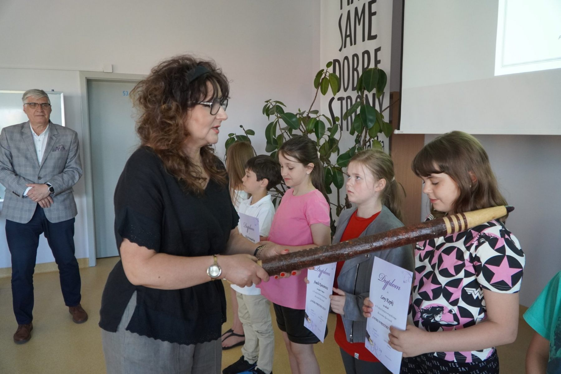 Pani Anna Markowska za pomocą dużego, drewnianego pióra pasuje uczniów na „Mistrza Ortografii”.