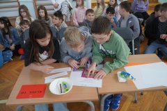 Troje Uczniów klasy 3d siedzi przy stoliku i dopasowuje zdjęcia zwierząt do zapisu kolejnych zagadek. Za Dziećmi siedzą pozostali Członkowie zespołu.