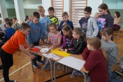 Trzech Uczniów klasy 3a siedzi przy stoliku i układa zdania z rozsypanek wyrazowych. Chłopcy są wspomagani przez stojących obok  pozostałych Członków zespołu.