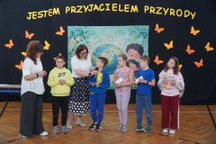 Pięcioro Trzecioklasistów prezentuje rozwiązania wylosowanych wcześniej zagadek przyrodniczych. Razem z dwiema  Nauczycielkami stoją na tle okolicznościowej dekoracji. 