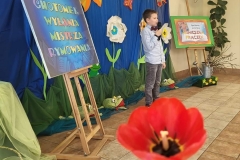 Dominik stoi na środku sceny i recytuje wiersz, w tle kolorowe wiosenne dekoracje.