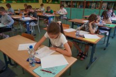 Sala lekcyjna w której uczniowie siedzą pojedynczo przy ławkach pisząc konkurs.