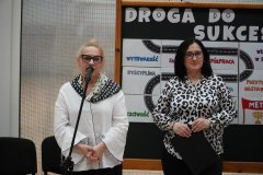 Dwoje dorosłych na apelu szkolnym, jedna osoba mówi do mikrofonu. W tle transparent z napisem „Droga do Sukcesu”.