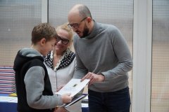 Mężczyzna wręcza książkę chłopcu, a kobieta patrzy z uśmiechem. Mężczyzna ma na sobie szary sweter, a chłopiec czarno-szarą kurtkę.