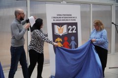 Trzy osoby dorosłe w scenerii wydarzenia odsłaniającej nowe logo szkoły pod nazwą „Szkoła Podstawowa z Oddziałami Integracyjnymi nr 22 im. Janusza Korczaka w Płocku”. Baner zawiera logo otwartej książki i ilustracje przedstawiające dzieci.