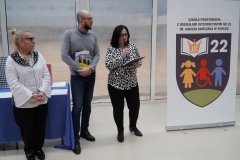 Trzy osoby stoją przy banerze reklamującym nowe logo szkoły. Kobieta w okularach trzyma mikrofon i przemawia, podczas gdy mężczyzna obok niej przegląda materiały. Trzecia kobieta stoi po lewej stronie, wszystko w dobrze oświetlonym pomieszczeniu.