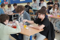 W sali lekcyjnej znajdują się grupy konkursowe, na pierwszym planie trzyosobowa grupka, która wykonuje doświadczenie według insrtukcji. Uczestnicy mają okulary ochronne oraz rękawiczki, a na ławkach znajdują się kubeczki z kolorowymi cieczami, bibuły, pipety, stojaki oraz pojemniki z cukrem.