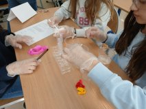 Zbliżenie na łwkę szkolną, przy której znajdują się trzy dziewczynki- uczestniczki konkursu. Mają okulary ochronne oraz rękawiczki, a na stoliczku znajdują się: kubeczki z kolorowymi cieczami, bibuły, pipety, stojak oraz pojemnik z cukrem.