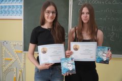 W sali lekcyjnej na tle zielonej tablicy szkolnej znajdują się dwie uczestniczki konkursu- zdobywczynie 2 miejsca w konkursie. Uczennice trzymają w rękach tematyczne dyplomy i nagrody książkowe.
