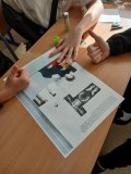 Zbliżenie na szkolną łąwkę, na której troje uczniów wypełnia konkursową kartę pracy dotyczącą parcy Marii Skłodowskiej- Curie; na karcie nawiązanie do prześwietleń RTG. Uczestnicy ukazują swoje zadowolenie poprzez pokazanie kciuków uniesionych w górę.