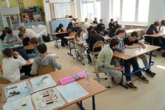 Sala lekcyjna, w której odbywa się konkurs, kilka grup uczniów. Na pierwszym planie znajduje się ławka szkolna, na której złożone są wypełnione przez uczestników konkursu karty pracy. W tle dygestrium chemiczne.