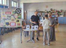 Pani dyrektor wręcza nagrodę dziecku. Obok tablica z wydrukowanymi pracami z konkursu multimedialnego. W tle biblioteka.