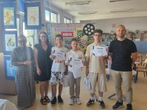 Pani dyrektor,nauczyciele oraz dzieci pozują do zdjęcia na tle tablicy z wydrukowanymi pracami z konkursu multimedialnego. W tle biblioteka.