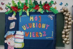 Zdjęcie przedstawia dekorację wykonaną w związku ze Szkolnym Konkursem Recytatorskim. Na szafirowym tle znajduje się kolorowy napis „Mój ulubiony wiersz”. Z jego lewej strony widnieje sylweta chłopca w birecie, który trzyma w rękach plik książek. Dookoła przywieszone są świąteczne dekoracje- girlandy bombek i kwiatów ostrokrzewu.