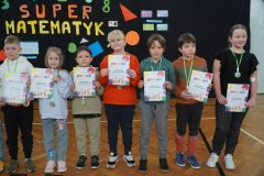 Na tle dekoracji konkursowej stoi siedmioro dzieci. Mają dyplomy oraz medale za udział w konkursie Super Matematyk.