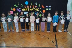Dwunastu uczniów stoi na parkiecie sali gimnastycznej i pokazuje swoje dyplomy i medale, które otrzymali za udział w Super Matematyku.