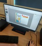 Zdjęcie przedstawia monitor komputera z otwartym programem do edycji, prawdopodobnie do tworzenia okładki książki. Na ekranie widoczna jest okładka książki "Król Maciuś Pierwszy" oraz tekst "Twórczość" (w języku polskim). W prawym dolnym rogu widać dłoń trzymającą myszkę komputerową.