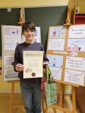 Zdjęcie uczennicy w okularach, trzymającej dyplom za przygotowanie prezentacji multimedialnej "Janusz Korczak w sercach dzieci!". Stoi obok tablicy z cytatami i rysunkami na temat Korczaka. Trzyma również torbę z nagrodami.