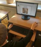 Ujęcie zza pleców ucznia siedzącego przy komputerze. Na monitorze widać okładkę lub slajd prezentacji z portretem Janusza Korczaka i tekstem: "Janusz Korczak. Patron naszej szkoły".