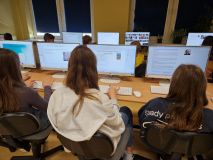 Zdjęcie z tyłu przedstawia grupę uczniów, prawdopodobnie w pracowni komputerowej, siedzących przy biurkach i pracujących na komputerach. Monitory pokazują różne informacje i teksty związane z twórczością Janusza Korczaka.