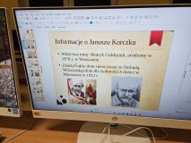 Zdjęcie monitora komputera z otwartą prezentacją (prawdopodobnie w programie LibreOffice Impress). Slajd zawiera tytuł "Informacje o Januszu Korczaku" oraz fakty z jego życia, w tym jego właściwe imię (Henryk Goldszmit) i rok założenia domu sierot. Poniżej znajdują się dwa archiwalne zdjęcia Janusza Korczaka.
