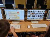 Ujęcie zza pleców dwojga uczniów pracujących przy komputerach. Na lewym monitorze widać slajd prezentacji z cytatem: "Wszystko co osiągnięte rozumem, naciskiem, przemocą, jest nietrwałe". Na prawym monitorze widoczna jest galeria zdjęć Janusza Korczaka.