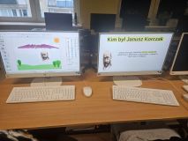 Zdjęcie dwóch monitorów komputerowych ustawionych na biurku. Na obu ekranach widać informacje o Januszu Korczaku. Prawy monitor wyświetla pytanie: "Kim był Janusz Korczak" i krótką odpowiedź, w której wymienione jest jego właściwe imię (Henryk Goldszmit), data i miejsce urodzenia oraz data śmierci (w Treblince).