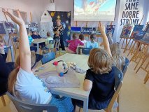 uczniowie siedzą przy stołach, kilka osób ma podniesione ręce i zgłasza się do odpowiedzi, w tle wystawa zdjęć o locie Sławosza oraz prowadzący zajęcia nauczyciel