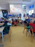 uczniowie siedzą przy stolikach i oglądają film o Sławoszu Uznańskim-Wiśniewskim, w tle wystawa zdjęć, papierowa rakieta i „ścianka” przedstawiająca niebo z planetami i gwiazdami