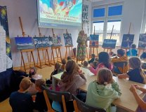 uczniowie siedzą przy stołach i słuchają prowadzącego nauczyciela, który opowiada o wystawie zdjęć zgromadzonych w bibliotece