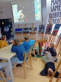 uczniowie siedzą przy stołach, trzy osoby leżą na dywanikach, wszyscy oglądają film, na ekranie postać astronauty w białym hełmie